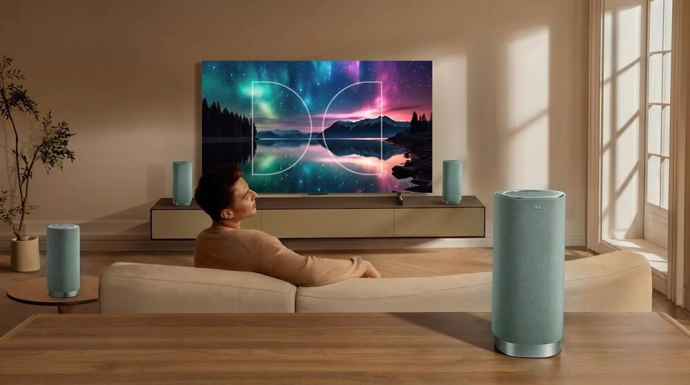 W salonie znajduje się duży telewizor wyświetlający kolorowy krajobraz z jeziorem i drzewami. Na pierwszym planie widoczna jest kolumna głośnikowa w jasnym kolorze.
