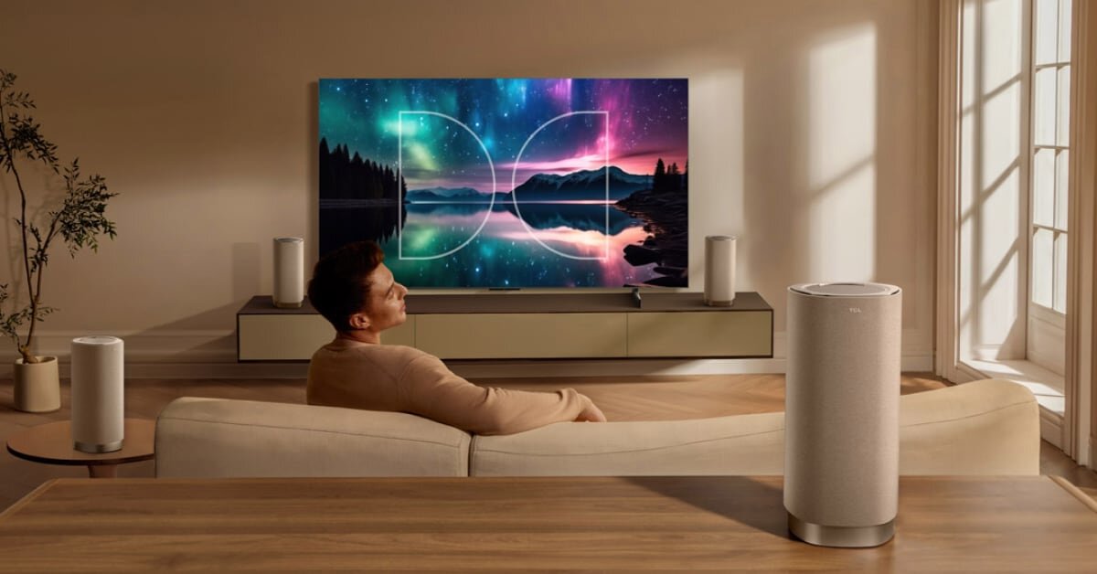 W salonie znajduje się duży telewizor wyświetlający kolorowy krajobraz z jeziorem i drzewami. Na pierwszym planie widoczna jest kolumna głośnikowa w jasnym kolorze.