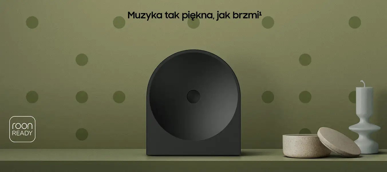 2026-hw-ls50h-kv-pc 1352x600-2 Na obrazie znajduje się czarny głośnik o nowoczesnym designie na tle zielonej ściany z kropkami. Widoczny jest tekst: \'Muzyka tak piękna, jak brzmi¹\' oraz logo \'roon READY\'.