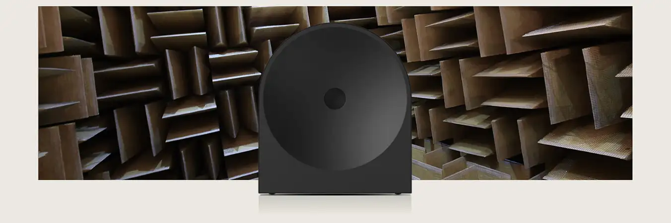 2026-hw-ls50h-samsung-audio-lab-pc 1352x450 Czarny głośnik o okrągłym kształcie umieszczony na tle pomieszczenia z panelami akustycznymi. Panele mają kształt prostokątnych, wystających elementów w kolorze brązowym.