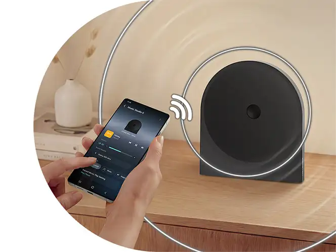 2026-hw-ls50h-top-4-wireless-music-streaming-mo 664x498 Osoba trzyma smartfon z aplikacją do odtwarzania muzyki, obok stoi czarny głośnik na drewnianej powierzchni. Wokół głośnika widoczne są graficzne fale dźwiękowe.