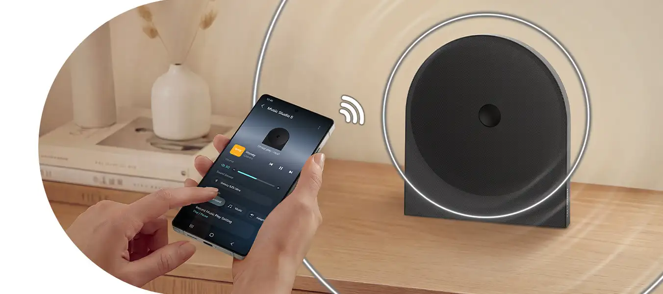 2026-hw-ls50h-top-4-wireless-music-streaming-pc 1352x600 Osoba trzyma smartfon z aplikacją \'Music Studio 5\', obok stoi czarny głośnik z widocznym symbolem Wi-Fi. W tle znajduje się wazon i książki na drewnianej półce.