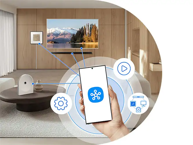 2026-hw-ls51h-smartthings-app 664x498 Wnętrze nowoczesnego salonu z telewizorem na ścianie, na którym wyświetlany jest krajobraz górski. Na pierwszym planie ręka trzymająca smartfon z ikoną aplikacji, wokół której znajdują się symbole ustawień, odtwarzania i połączeń.