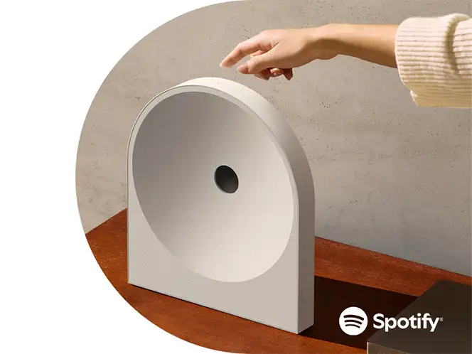 2026-hw-ls51h-spotify-tap 664x498 Biały głośnik o nowoczesnym, zaokrąglonym kształcie stoi na drewnianej powierzchni, a nad nim unosi się ręka. W prawym dolnym rogu widoczne jest logo Spotify.
