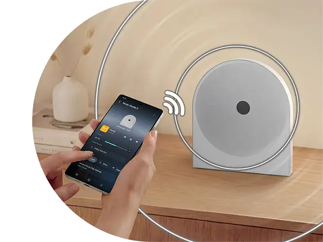 2026-hw-ls51h-top-4-wireless-music-streaming-mo 664x498 Na drewnianej powierzchni stoi biały głośnik o okrągłym kształcie. Obok osoba trzyma smartfon z aplikacją muzyczną na ekranie.