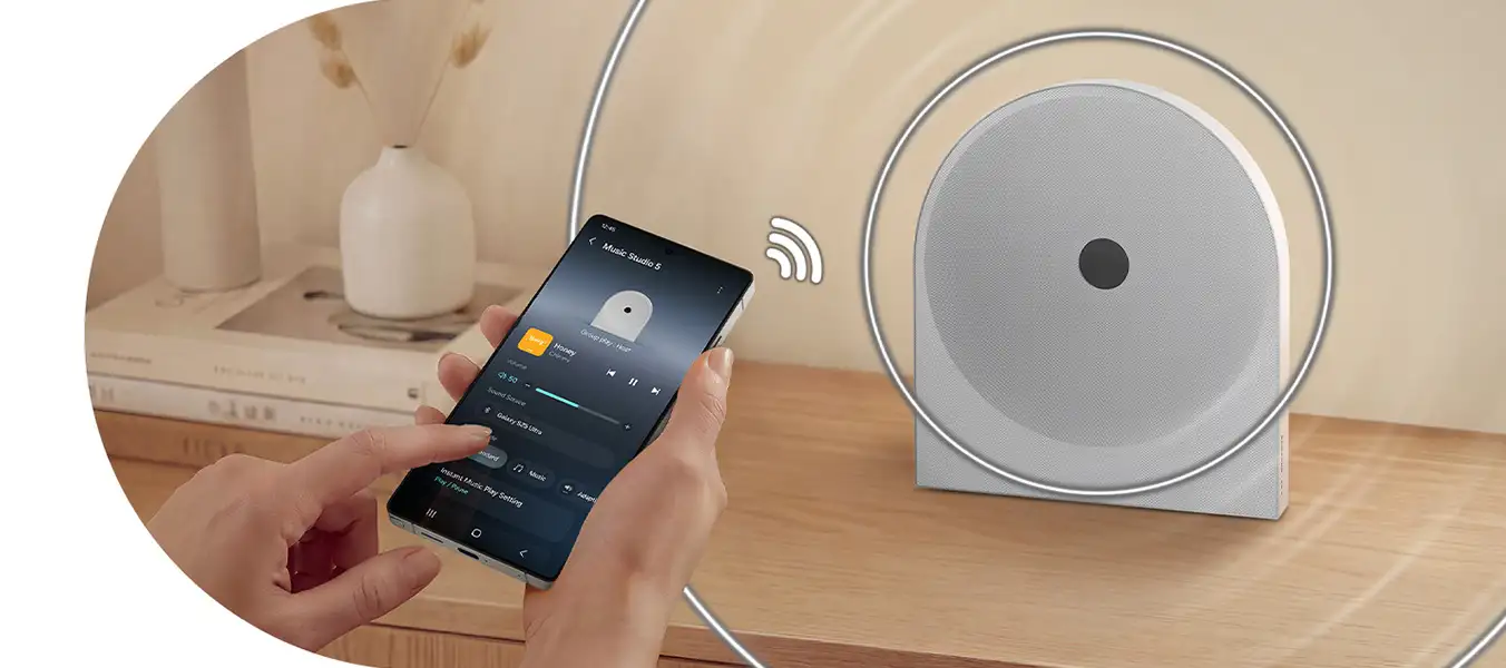 2026-hw-ls51h-top-4-wireless-music-streaming-pc 1352x600 Na drewnianym blacie stoi biały głośnik o okrągłym kształcie z czarnym punktem na środku. Obok osoba trzyma smartfon z aplikacją muzyczną, na ekranie widoczny jest tekst \'Music Studio 5\'.