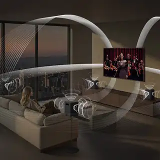 2026-hw-ls70-top-4-q-symphony 320x320 Osoba siedzi na kanapie w salonie, oglądając telewizor z wyświetlanym koncertem. Wokół niej widoczne są graficzne fale dźwiękowe, sugerujące przestrzenny dźwięk z głośników.