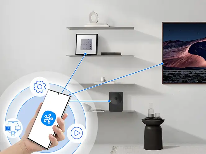 2026-hw-ls70h-smartthings-app 664x498 Na obrazie widać rękę trzymającą smartfon z ikoną aplikacji, a w tle znajdują się półki z dekoracjami oraz telewizor wyświetlający krajobraz. Na ekranie smartfona widoczne są ikony ustawień i odtwarzania.