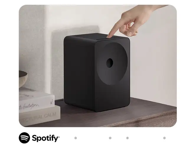 2026-hw-ls70h-spotify-tap 664x498 Czarny głośnik o prostokątnym kształcie stoi na drewnianej powierzchni, obok książek. Nad głośnikiem widoczna jest dłoń wskazująca na jego górną część, a na dole znajduje się logo Spotify.