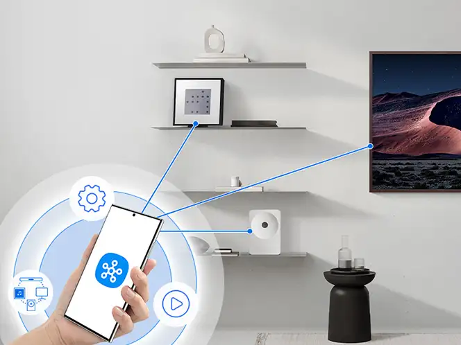 2026-hw-ls71h-smartthings-app 664x498 Na obrazie widać wnętrze pokoju z półkami na ścianie, na których znajdują się różne przedmioty, oraz telefon komórkowy z ikonami na ekranie. Na ścianie wisi obraz przedstawiający krajobraz, a na stoliku stoi butelka i szklanka.