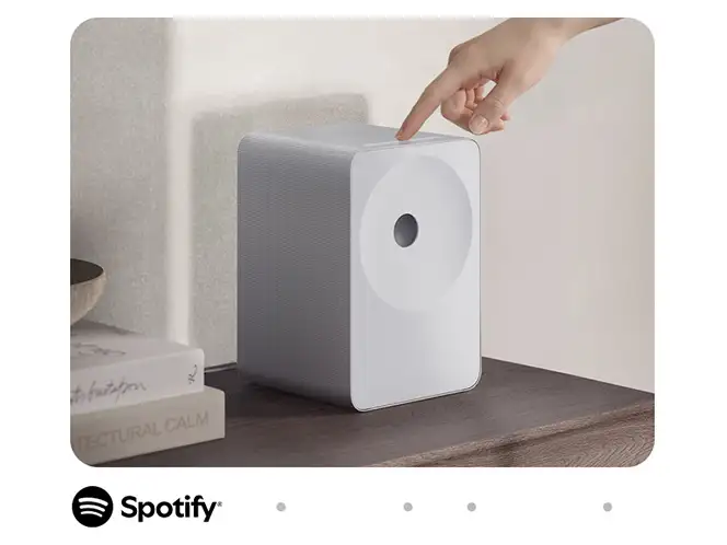 2026-hw-ls71h-spotify-tap 664x498 Biały głośnik o prostokątnym kształcie stoi na drewnianej powierzchni, obok książek. Nad głośnikiem widoczna jest ręka wskazująca na jego górną część, a na dole znajduje się logo Spotify.