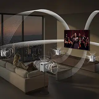 2026-hw-ls71h-top-4-q-symphony 320x320 W salonie znajduje się telewizor wyświetlający koncert, a wokół niego rozmieszczone są głośniki emitujące fale dźwiękowe. Na kanapie siedzi osoba, a przez okno widać panoramę miasta o zmierzchu.