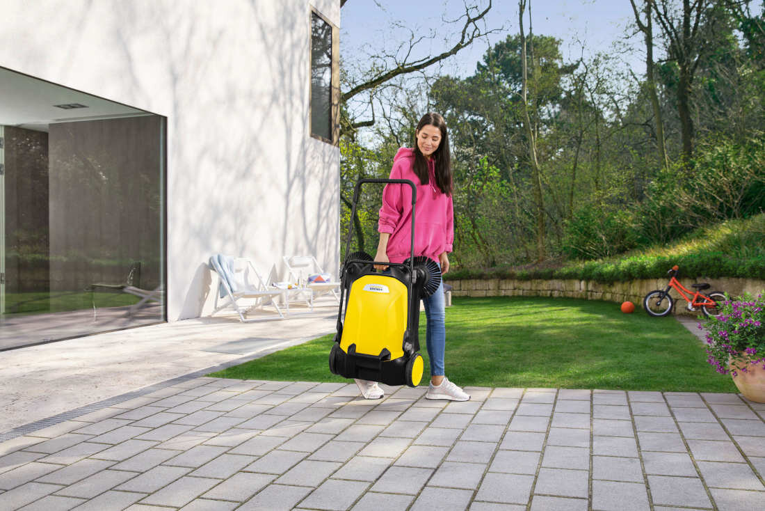 Osoba trzyma żółtą zamiatarkę z widocznym logo KARCHER na tle ogrodu z trawnikiem i rowerkiem dziecięcym. W tle znajduje się nowoczesny budynek z dużymi oknami.