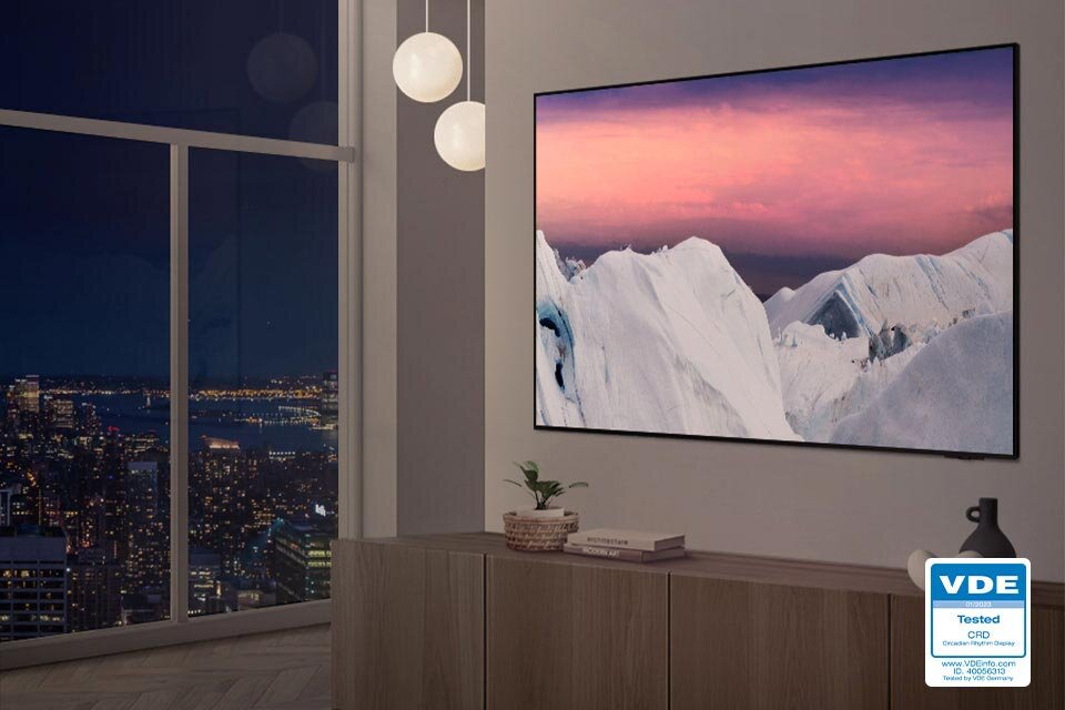 W nowoczesnym wnętrzu na ścianie wisi duży telewizor wyświetlający obraz gór lodowych o zachodzie słońca. Obok telewizora znajduje się certyfikat z napisem \'VDE Tested CPR\'.