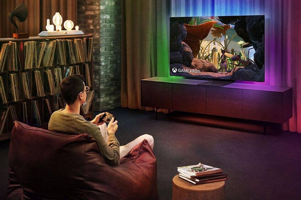 Osoba siedzi na kanapie i gra na konsoli, patrząc na duży telewizor wyświetlający kolorową grafikę z napisem \'GAME PASS\'. W tle widać regał z płytami i dekoracyjne oświetlenie.