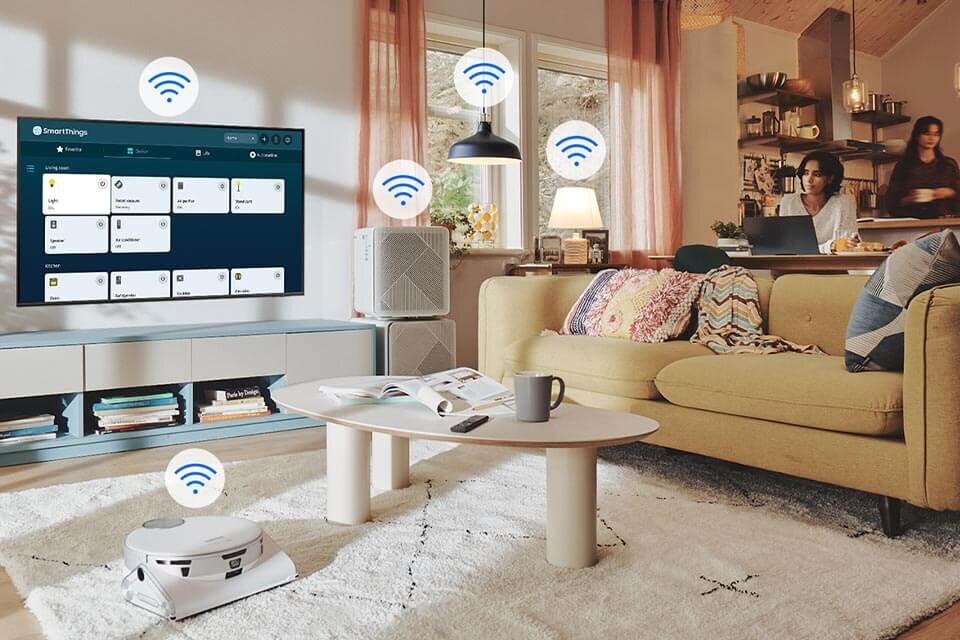W salonie znajduje się telewizor z wyświetlonym interfejsem \'SmartThings\', obok którego widoczne są ikony sygnału Wi-Fi. Na dywanie stoi robot sprzątający, a w tle widać sofę, stolik kawowy i osoby przy komputerze.