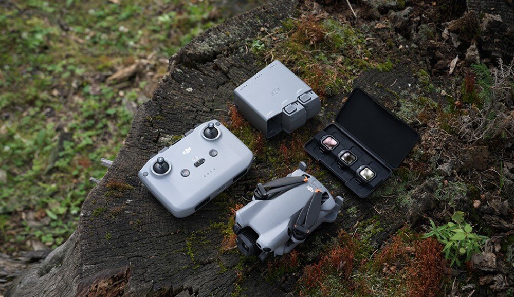 Na pniu drzewa leży szary dron z złożonymi ramionami, obok niego znajduje się kontroler, bateria oraz otwarte etui z trzema filtrami. W tle widoczna jest trawa i mech.