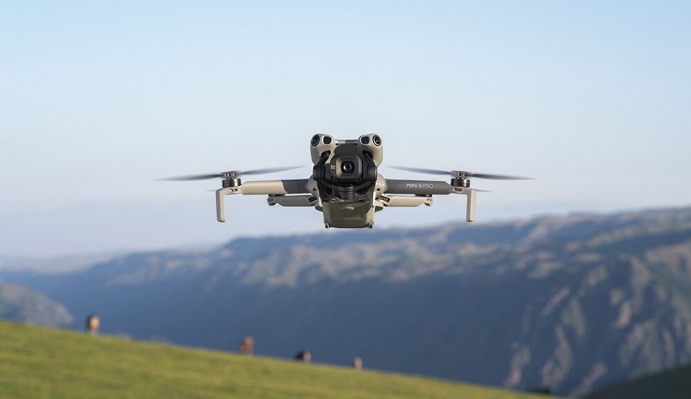Dron w locie nad zielonym wzgórzem z rozmytym tłem górskim. Na dronie widoczny jest napis \'MINI 5 PRO\'.