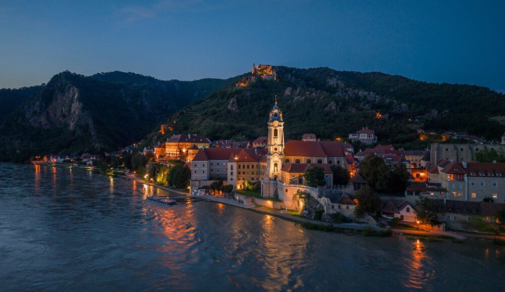 Widok na malownicze miasteczko nad rzeką o zmierzchu, z oświetlonymi budynkami i kościołem na pierwszym planie. W tle widać wzgórza i zamek na szczycie.