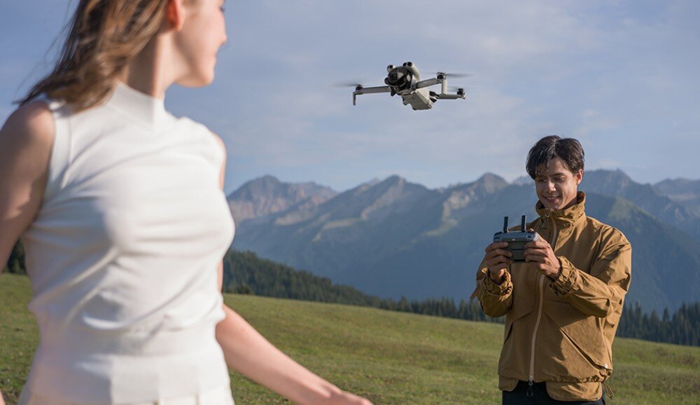 Na tle górskiego krajobrazu widać drona w locie oraz osobę trzymającą kontroler. Obok stoi inna osoba w białej bluzce.