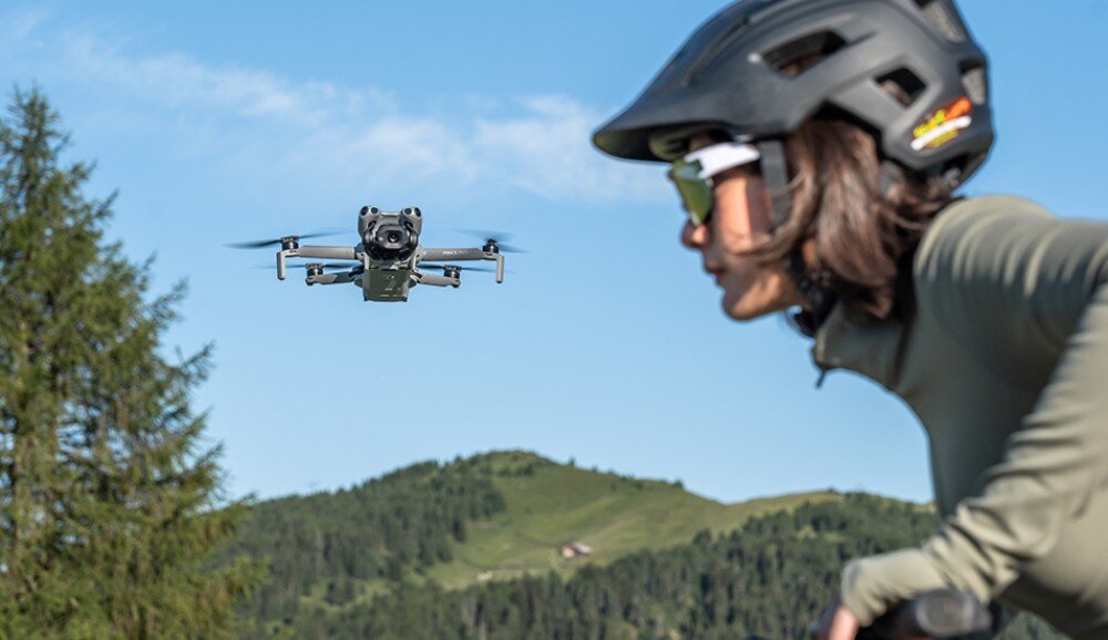 Dron unosi się w powietrzu na tle zielonych wzgórz i drzew. Obok znajduje się osoba w kasku i okularach przeciwsłonecznych.