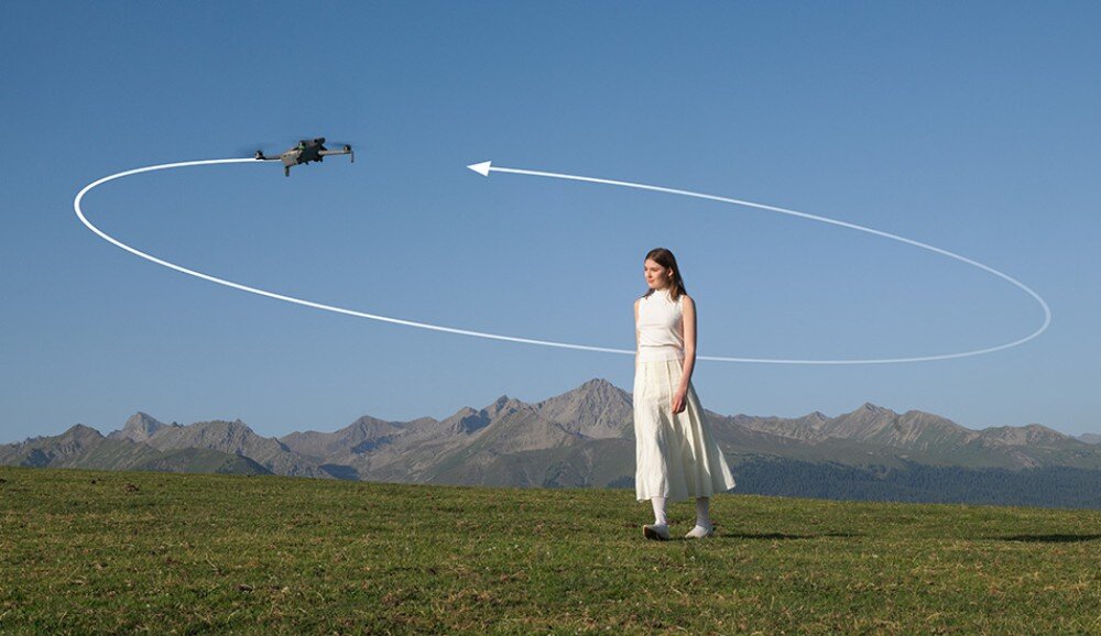 Na tle górskiego krajobrazu widać drona unoszącego się w powietrzu oraz osobę stojącą na trawie. Dron jest otoczony białą linią z strzałką, sugerującą jego trasę lotu.