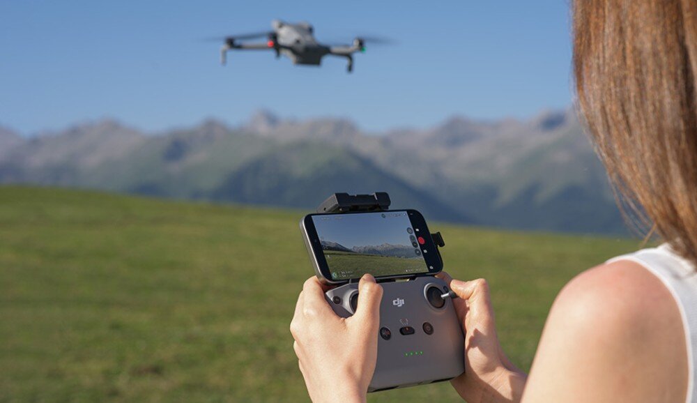 Osoba trzyma kontroler z ekranem, na którym widać obraz z kamery drona unoszącego się w tle na tle górskiego krajobrazu. Na kontrolerze widoczne jest logo \'dji\'.