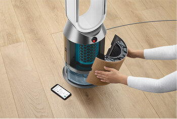 Osoba wymienia filtr w oczyszczaczu powietrza z wentylatorem, obok leży smartfon z wyświetlonym interfejsem aplikacji. Urządzenie stoi na drewnianej podłodze.