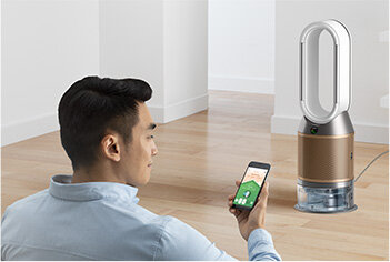 Osoba siedzi na podłodze, trzymając smartfon z aplikacją, przed oczyszczaczem powietrza z wentylatorem w nowoczesnym wnętrzu. Oczyszczacz ma cylindryczny kształt z dużym, owalnym otworem na górze.