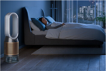 W sypialni znajduje się oczyszczacz powietrza z wentylatorem o nowoczesnym designie, obok łóżka, na którym śpi osoba. W tle widoczne jest duże okno z widokiem na miasto nocą.