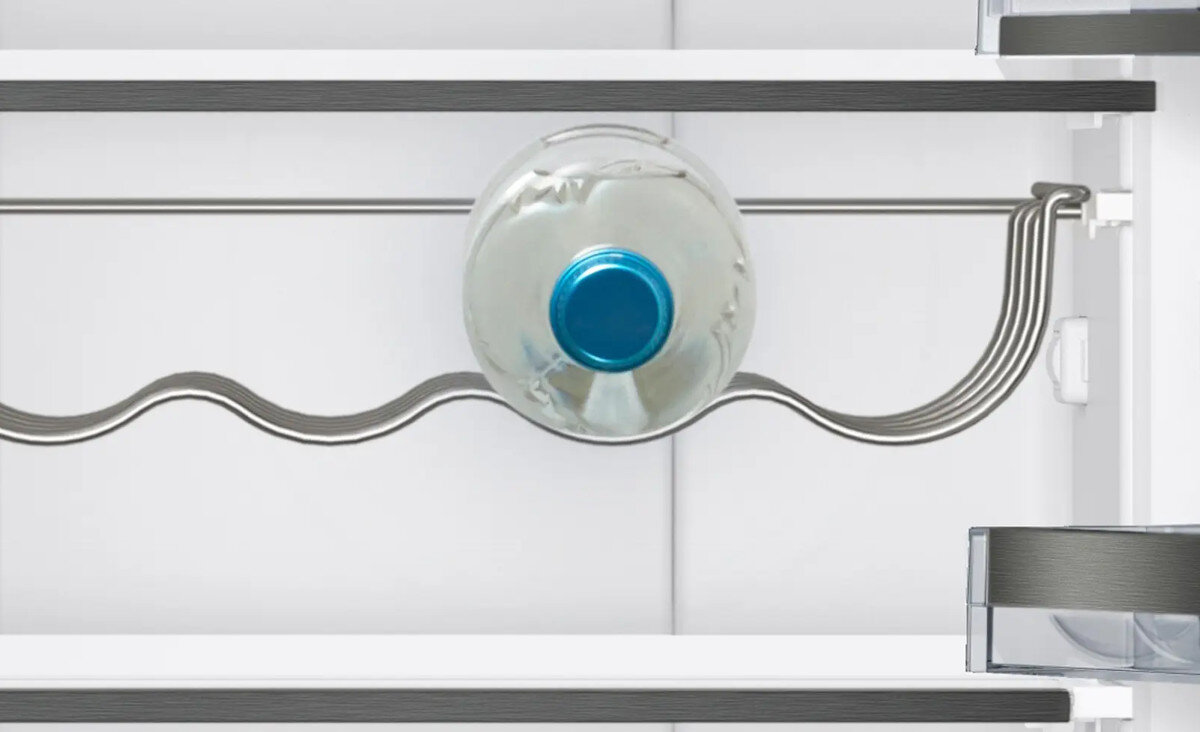 Wnętrze lodówki z widoczną półką na butelki, na której znajduje się jedna plastikowa butelka z niebieskim korkiem. Półka ma falisty kształt, a wnętrze lodówki jest białe z szarymi elementami.