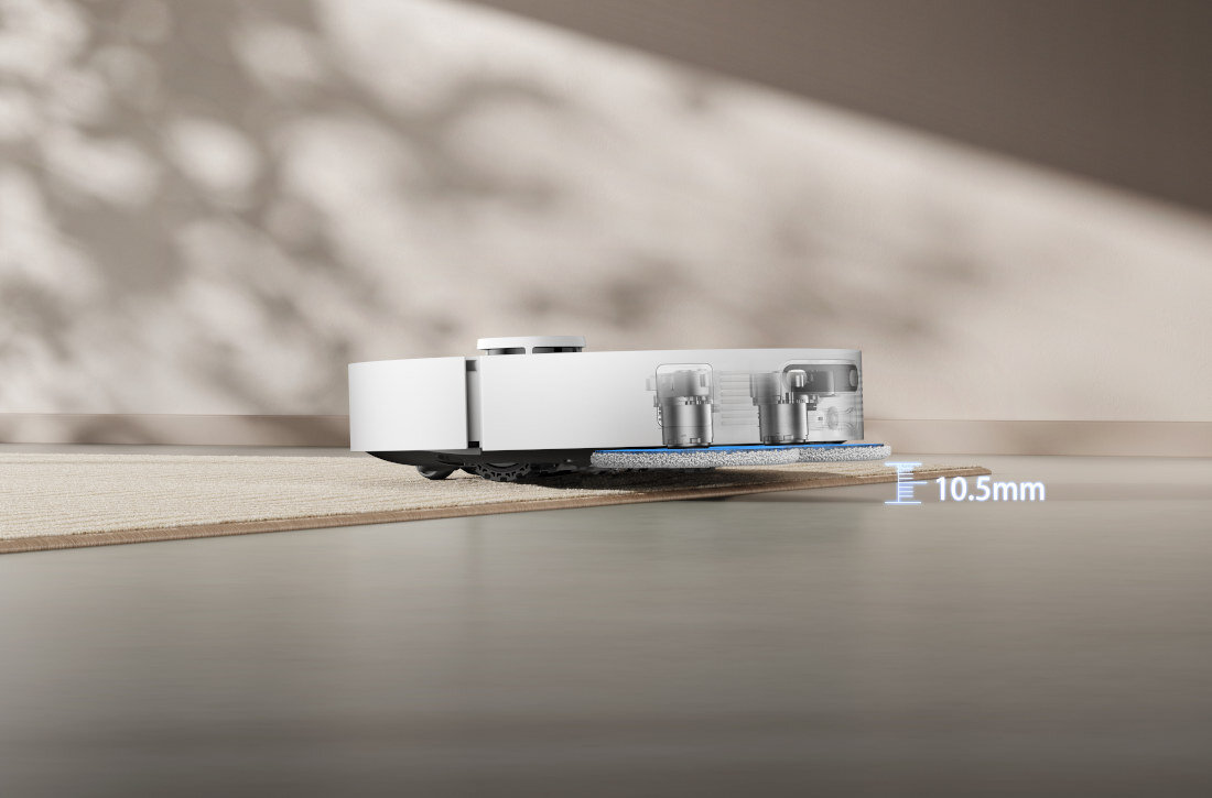 Robot sprzątający w kolorze białym pokonuje próg o wysokości 10,5 mm. W tle widoczne są rozmyte cienie na ścianie.
