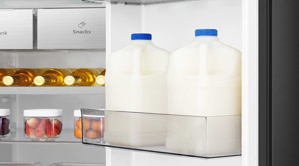 Wnętrze lodówki z dwoma dużymi pojemnikami na mleko na drzwiach oraz półką z butelkami i słoikami z owocami. Widoczne są napisy \'Snacks\' i \'Drink\'.