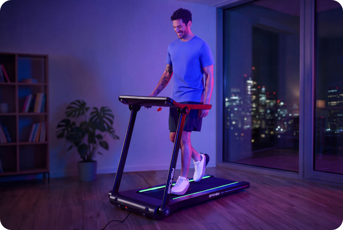 Mężczyzna ćwiczy na bieżni elektrycznej w nowoczesnym wnętrzu z dużymi oknami. Na bieżni widoczne jest logo \'SPOKEY\'.