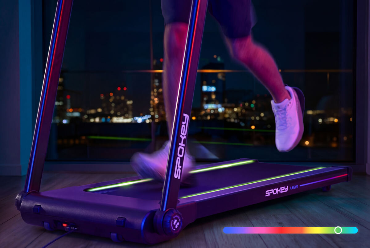 Osoba biegnie na bieżni elektrycznej marki SPOKEY w oświetlonym pomieszczeniu z widokiem na miasto nocą. Na bieżni widoczne są napisy \'SPOKEY\' i \'Lean\'.