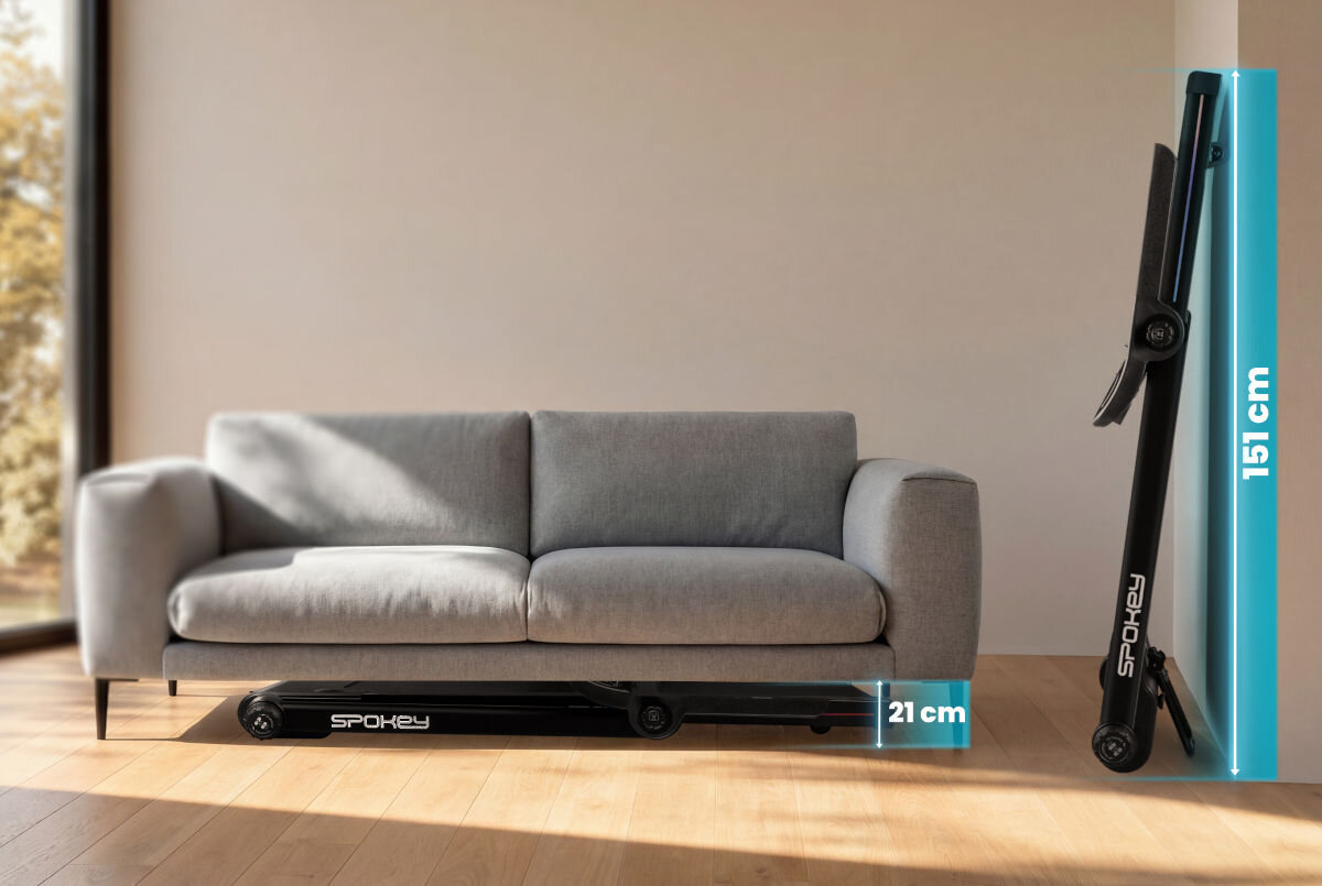 Na obrazie znajduje się szara sofa, przed którą leży złożona bieżnia z widocznym logo \'SPOKEY\'. Obok bieżni widoczne są wymiary: 21 cm w pozycji poziomej i 151 cm w pozycji pionowej.
