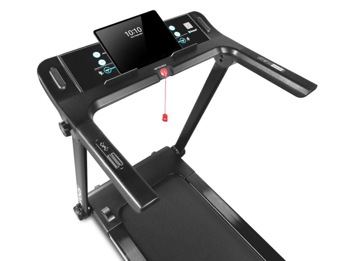Bieżnia elektryczna z czarnym panelem sterującym, na którym znajduje się ekran z wyświetlaczem pokazującym godzinę 10:10. Na panelu widoczne są przyciski funkcyjne oraz czerwony klucz bezpieczeństwa.