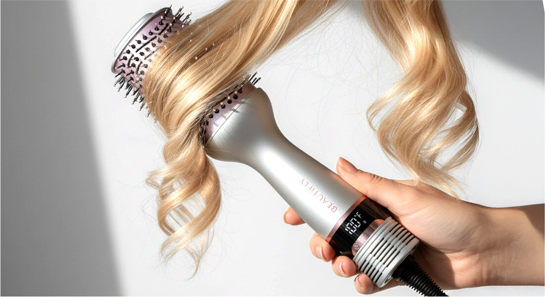 Osoba trzyma suszarko-lokówkę, która stylizuje blond włosy. Na urządzeniu widoczny jest wyświetlacz z temperaturą oraz napis \'BEAUTIFLY\'.