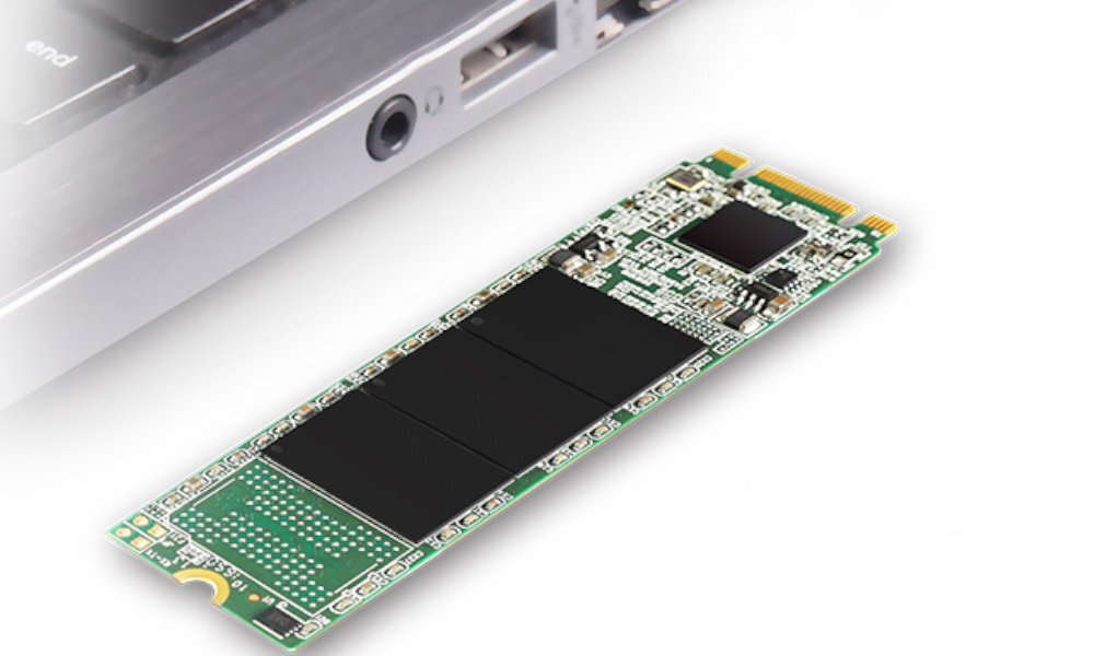 Na obrazie widoczny jest zielony dysk SSD z czarnymi elementami, leżący obok fragmentu laptopa. Dysk ma prostokątny kształt i widoczne są na nim różne komponenty elektroniczne.