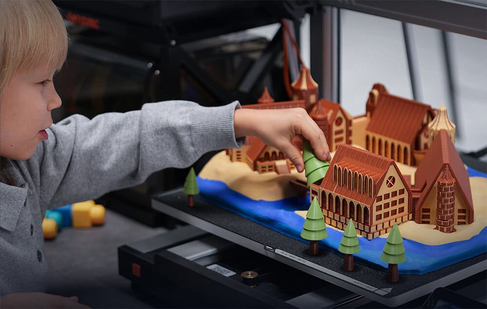 Drukarka 3D ANYCUBIC Kobra 3 Max Combo Wi-Fi, LAN + Dysza + Filament PLA Czarny Dziecko układające elementy wielokolorowej, wydrukowanej w 3D makiety miasteczka, która znajduje się na platformie roboczej drukarki 3D systemy kompensacji rezonansu dynamiczna kontrola przepływu redukcja wibracji łożyska SG15 przeciwdziałanie przesunięcia warstw stabilność podwójna śruba osi Z