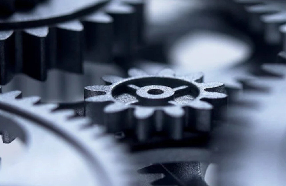 Zbliżenie na metalowe zębatki, które są ze sobą połączone i tworzą mechanizm. Zębatki mają różne rozmiary i są w odcieniach szarości.