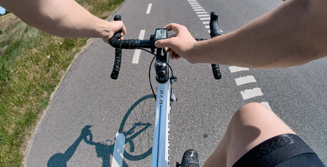 Osoba jedzie na rowerze po asfaltowej drodze, trzymając kierownicę z zamontowanym licznikiem rowerowym. Widoczny jest fragment roweru oraz cień rowerzysty na drodze.