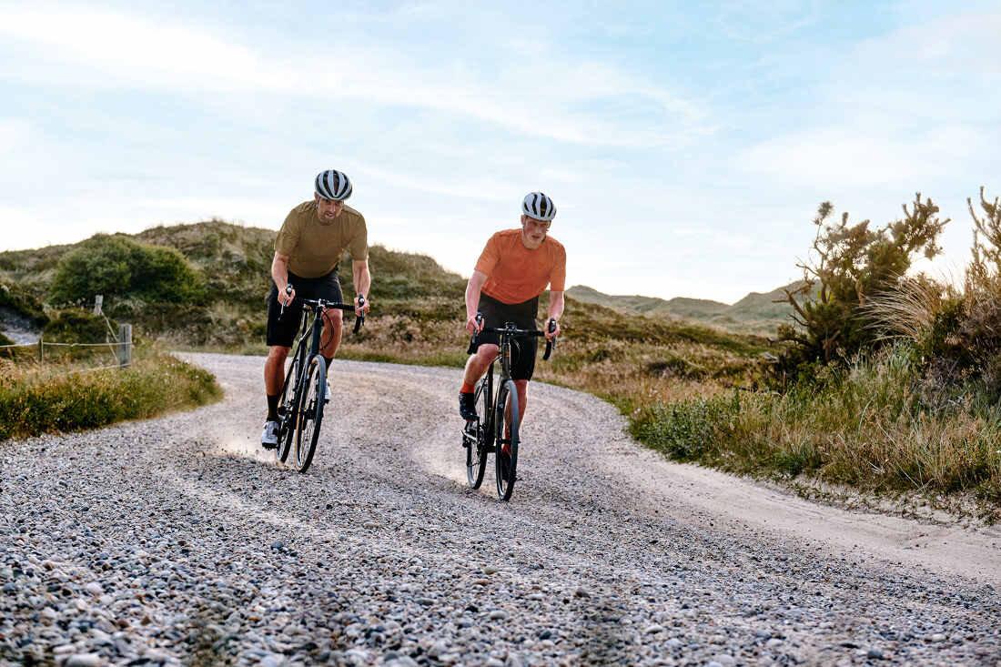 Dwóch rowerzystów jedzie po żwirowej drodze w otoczeniu zielonych wzgórz i krzewów. Obaj mają na sobie kaski i sportowe ubrania.