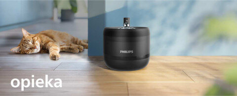 Na drewnianej podłodze stoi czarna fontanna z widocznym logo PHILIPS, obok leży śpiący kot. W lewym dolnym rogu znajduje się napis \'opieka\'.