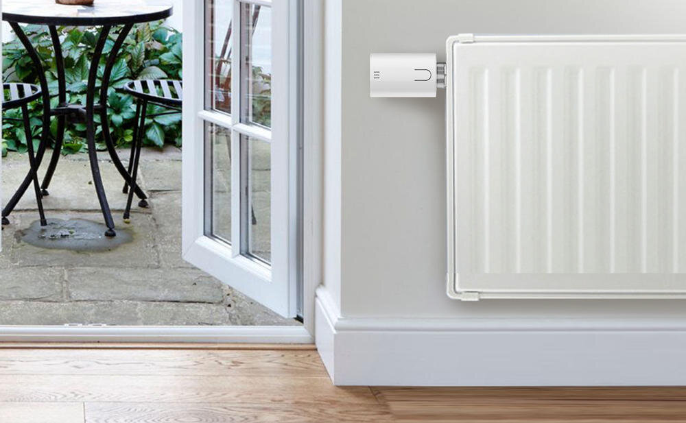 Na obrazie widać biały grzejnik z zamontowaną głowicą termostatyczną. W tle znajduje się otwarte drzwi prowadzące na taras z widocznym stolikiem i krzesłem.