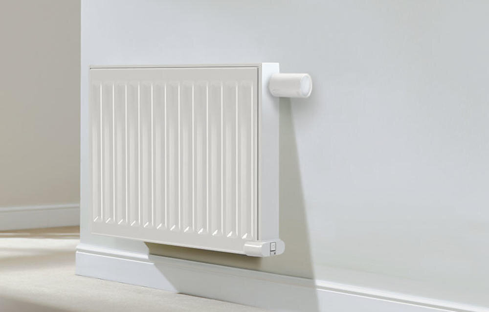 Na obrazie widoczny jest biały grzejnik zamontowany na ścianie. Obok grzejnika znajduje się biała głowica termostatyczna.