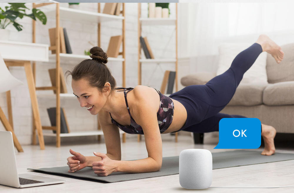 Osoba ćwiczy na macie przed laptopem w jasnym wnętrzu z półkami. Obok znajduje się biały głośnik z niebieską chmurką tekstową zawierającą napis \'OK\'.