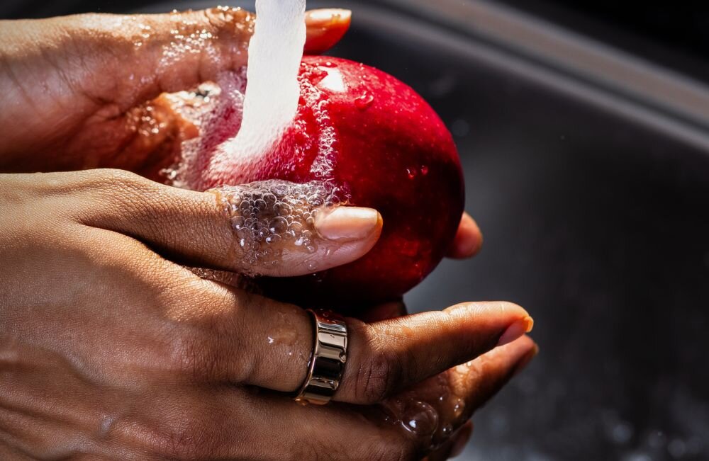 Osoba myje czerwone jabłko pod bieżącą wodą, na palcu widoczny jest złoty pierścień.