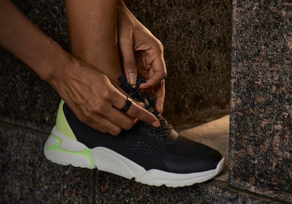 Osoba wiąże sznurowadła czarnego buta sportowego z białą podeszwą i zielonym akcentem. Na palcu widoczny jest ciemny pierścień.