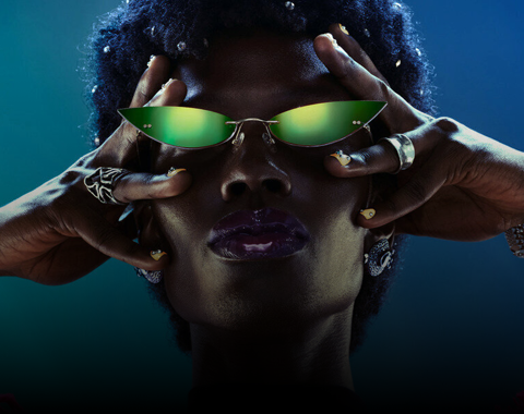 Osoba nosi zielone okulary przeciwsłoneczne o nietypowym kształcie. Na palcach widoczne są pierścionki, a tło jest ciemne.
